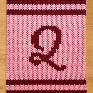 Puede incluir: Un artículo rectangular tejido con una gran letra "Q" burdeos sobre un fondo rosa. Los bordes superior e inferior presentan rayas burdeos, y la textura general sugiere una sensación acogedora y hecha a mano.