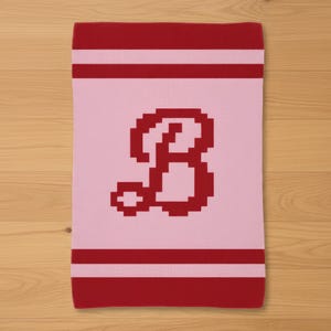 May include: A rectangular, knitted baby blanket in pink with a large red letter "B" in the center. The blanket has red stripes at the top and bottom. The blanket is made of soft material.