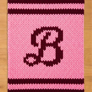 May include: A knitted, square-shaped item with a pink background and a large, dark burgundy letter "B" in the center. The top and bottom edges feature dark burgundy stripes, and the item is made of thick yarn.