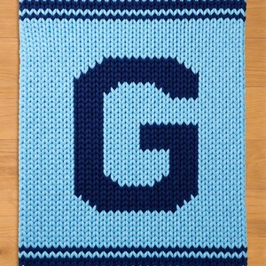 May include: A knitted blanket in light blue with a large, dark blue letter "G" in the centre. The blanket has dark blue stripes at the top and bottom edges, creating a contrasting border. The chunky knit texture is visible.