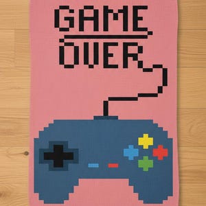May include: A pink blanket with a pixelated design featuring a blue video game controller and the words "GAME OVER" in black. The controller has colorful buttons and a black cord.