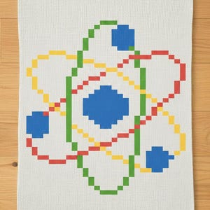 May include: White kitchen towel featuring a pixelated atom design in green, red, yellow, and blue. The atom graphic is centered on the towel, with a textured appearance. The towel is made of a soft material.