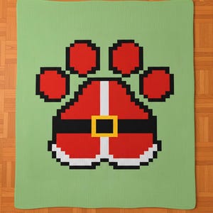 May include: A green blanket featuring a pixelated design of a Santa Claus-themed paw print. The paw print is red with black and white accents, including a black belt with a gold buckle. The blanket is rectangular.
