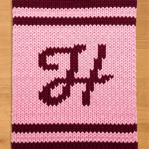 Könnte beinhalten: Eine rechteckige Strickdecke in Rosa- und Burgundertönen. Die Decke zeigt ein großes, stilisiertes Buchstabendesign in Burgunderrot auf rosa Hintergrund. Die oberen und unteren Kanten sind burgunderfarben, mit rosa und burgunderfarbenen Streifen.