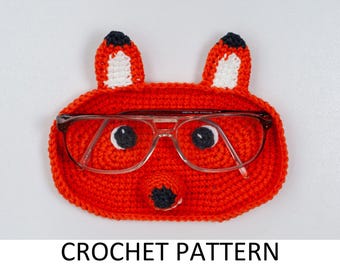 May include: A crocheted orange fox-shaped eyeglass holder with black and white accents. The fox has two ears, black eyes, and a black nose. Clear-framed eyeglasses rest inside the holder. The text "CROCHET PATTERN" is at the bottom.
