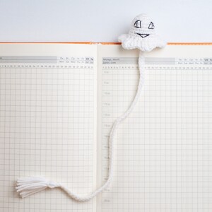 May include: A white crocheted ghost bookmark with a smiling face and a long tail. The bookmark is placed in a blank, orange-bound planner with a grid pattern.