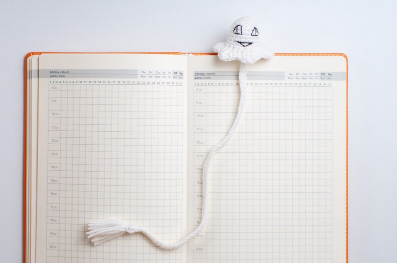 May include: A white crocheted ghost bookmark with a smiling face and a long tail. The bookmark is placed in a blank, orange-bound planner with a grid pattern.