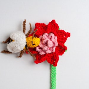 May include: A crocheted bee with white and yellow accents is perched on a red and pink crocheted flower with a green stem.