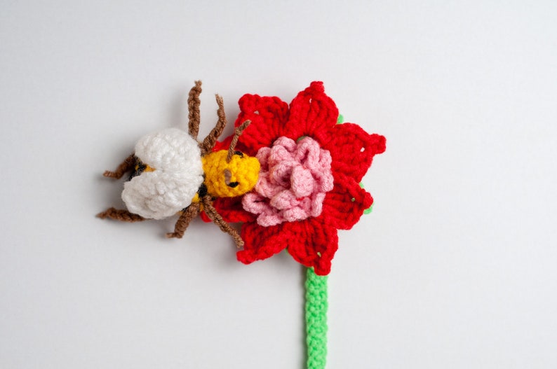 May include: A crocheted bee with white and yellow accents is perched on a red and pink crocheted flower with a green stem.