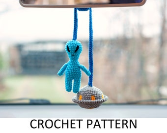 Puede incluir: Un extraterrestre azul de crochet con una nave espacial gris de crochet colgando de un espejo retrovisor. El extraterrestre tiene ojos negros y una expresión de enfado. CROCHET PATTERN está escrito en negro en la parte inferior de la imagen.