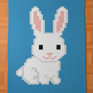Può includere: Una coperta blu con un motivo a coniglio bianco pixelato. Il coniglio ha orecchie interne rosa e un naso rosa. La coperta ha un aspetto strutturato. Il design è probabilmente per una cameretta.
