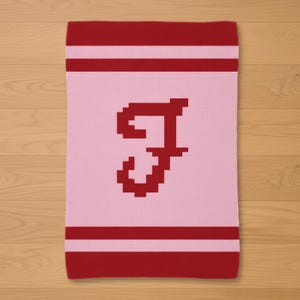 May include: A pink and red knitted blanket featuring the letter "J" in a pixelated design. The blanket has red stripes at the top and bottom edges. The background is a light pink color.