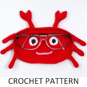 May include: Red crocheted crab-shaped glasses case with white eyes and a pink smile. The case has a pair of glasses inside. CROCHET PATTERN text is below the case.