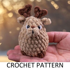 May include: A crocheted reindeer toy with brown antlers and a brown and beige body. The reindeer is sitting in the palm of a hand.