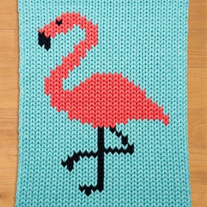 May include: A knitted square with a flamingo motif. The flamingo is depicted in coral and black against a light blue backdrop. The knitted texture is prominent, enhancing the design. The overall style is cheerful and decorative.