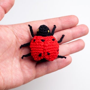 May include: A red and black crocheted ladybug with black legs and yellow eyes. The ladybug is sitting in the palm of a hand.