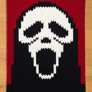 May include: A knitted rectangular throw blanket featuring a white skull design against a black background. The top and sides are framed with a red border. The skull has a wide open mouth.