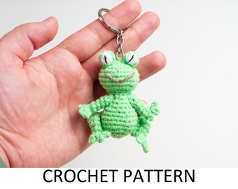 May include: Green crocheted frog keychain with white eyes and a brown smile. The frog is sitting on a hand.