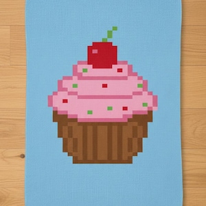 May include: A light blue kitchen towel featuring a pixelated cupcake design. The cupcake has pink frosting with green and red sprinkles, a brown base, and a red cherry on top. The towel is made of a soft material.