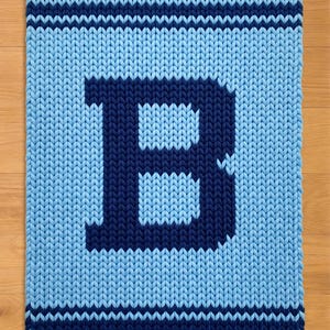 Può includere: Una coperta lavorata a maglia in tonalità di blu, con una grande lettera "B" in una tonalità di blu più scura. La coperta ha un aspetto strutturato con strisce orizzontali in alto e in basso, su una superficie di legno.