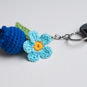 Op de afbeelding: Een blauwe gehaakte bloemen sleutelhanger met een groene stengel en een blauwe bloemknop. De sleutelhanger is bevestigd aan een zilveren sleutelring.
