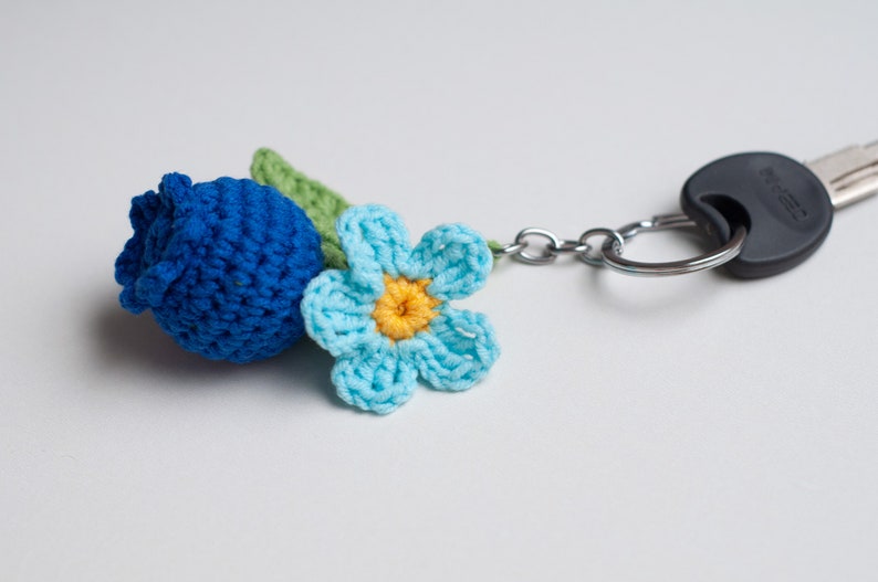 Op de afbeelding: Een blauwe gehaakte bloemen sleutelhanger met een groene stengel en een blauwe bloemknop. De sleutelhanger is bevestigd aan een zilveren sleutelring.
