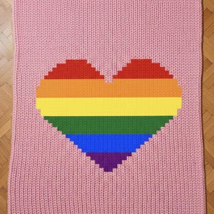 May include: A pink knitted blanket featuring a large heart design in the colors of the rainbow. The blanket is made of a soft, textured material and is displayed on a wooden floor.