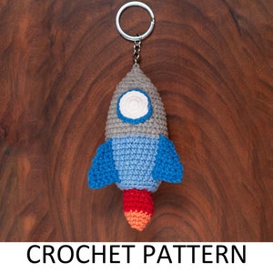 May include: A crocheted rocket ship keyring with a grey body, blue wings, and a red and orange flame. The keyring is attached to a silver ring.