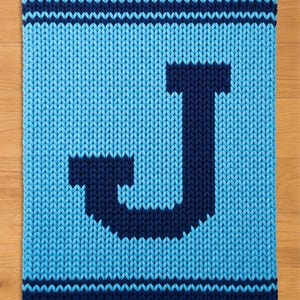 May include: A knitted rectangular blanket in shades of blue. The blanket features a large, dark blue letter "J" in the centre. The edges are bordered with dark blue stripes. The background is a lighter shade of blue.