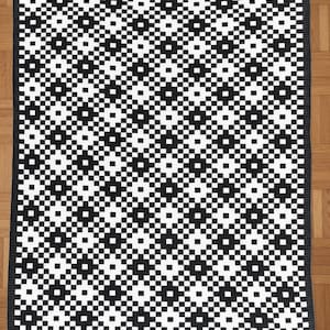 May include: A black and white geometric patterned blanket. The blanket features a repeating pattern of squares and diamonds in a high-contrast design. The edges are finished with a solid black border. The blanket is likely made of a soft, woven material.