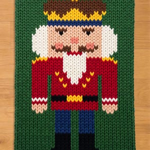 Könnte beinhalten: Ein gestrickter Weihnachtsteppich mit einem Nussknacker-Design. Der Nussknacker hat eine rote Jacke, blaue Hosen und eine braune und gelbe Krone. Der Hintergrund ist grün und der Teppich ist rechteckig.