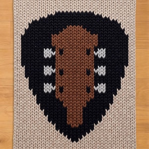 May include: A rectangular, knitted rug with a guitar design. The guitar is brown, set against a black heart shape, and surrounded by a beige border. The guitar has white accents. The rug is placed on a wooden surface.