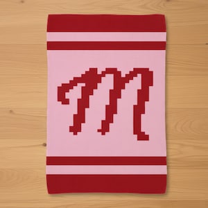 May include: A pink and red knitted blanket featuring a large, pixelated letter "M" in red. The blanket has red stripes at the top and bottom. The blanket is displayed on a wooden surface.