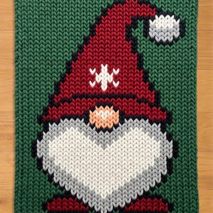 May include: A knitted Christmas gnome design on a green background. The gnome has a red hat with white snowflake accents, a white beard, and a peach-colored nose. The design is rectangular and appears to be a rug or decorative piece.