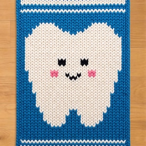 May include: A rectangular, knitted rug featuring a smiling tooth design. The tooth is white with pink cheeks and black eyes and a mouth. The rug has a blue border with white stripes at the top and bottom. The rug is made of a soft, textured material.
