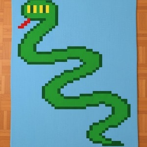 Op de afbeelding: Een gebreide deken met een gepixeld groen slangenontwerp op een lichtblauwe achtergrond. De slang heeft gele ogen en een rode tong. De deken is rechthoekig en lijkt handgemaakt.