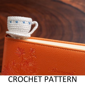 May include: A white crocheted teacup bookmark with blue accents, sitting on top of an orange leather-bound journal with the word "Bonnyeois" embossed on the cover.