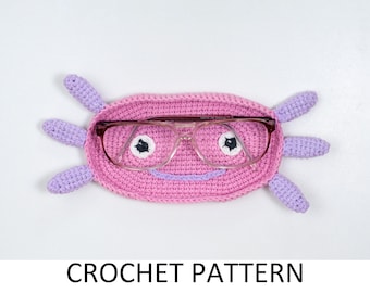 May include: A pink crocheted glasses holder shaped like an axolotl with purple accents. The holder has two black eyes and a smiling mouth. It is designed to hold eyeglasses.