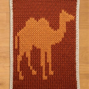 May include: A knitted camel design on a rectangular rug. The camel is in shades of orange and yellow against a brown background. The rug has a white border. This handmade item is suitable for a nursery or children's room.