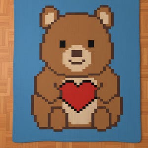 May include: A pixelated brown bear holding a red heart on a blue blanket. The bear has black eyes and a small smile. The blanket is square and appears to be knitted or crocheted, with a solid blue border.