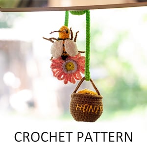 Pode incluir: Abelha e flor de crochê penduradas em um cordão verde com um pote de mel de crochê marrom que diz "Honey" pendurado abaixo.