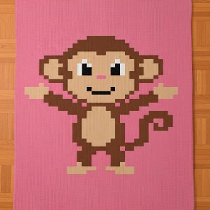 Puede incluir: Una manta rosa tejida con un diseño de mono pixelado. El mono tiene pelaje marrón, cara y vientre color beige, y está sobre un fondo rosa liso. Los brazos del mono están extendidos.