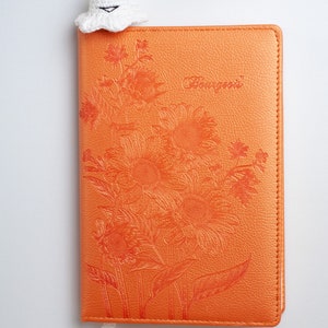 May include: A white crocheted ghost bookmark with a smiling face, attached to a white tassel, is placed on top of an orange leather journal with a floral design and the word "Bourgeois" embossed on the cover.