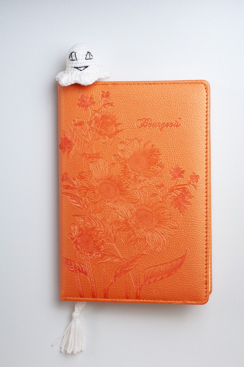 May include: A white crocheted ghost bookmark with a smiling face, attached to a white tassel, is placed on top of an orange leather journal with a floral design and the word "Bourgeois" embossed on the cover.