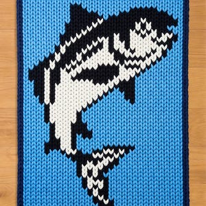 May include: A knitted blue blanket with a navy blue border. The blanket features a black and white orca whale design in the center. The whale is jumping out of the water. The blanket is made of a soft, textured material.