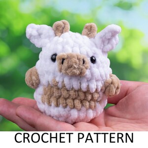 May include: A crocheted stuffed animal resembling a cow, with a white body and brown spots. The cow is sitting in a person's hand, with a green and white blurred background.