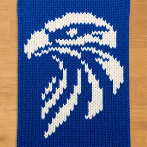 May include: A knitted blue and white decorative square with a stylized design. The design features a white abstract shape against a solid blue background. The item is made of a thick, textured yarn.