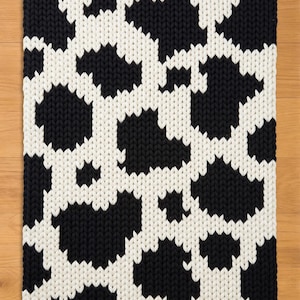 May include: A rectangular, hand-knitted rug with a cowhide pattern. The rug features large, irregular black shapes against a white background. The thick yarn creates a textured surface, suitable for a living room or bedroom.