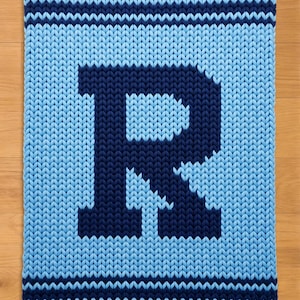 Peut inclure: Une couverture tricotée avec un grand "R" bleu marine sur un fond bleu clair. La couverture a un aspect texturé avec des rayures bleu marine en haut et en bas. Elle est présentée sur une surface en bois. Dimensions non visibles.