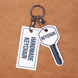 May include: A white keychain with a silver key ring and two tags. One tag says "HANDMADE KEYCHAIN" and the other tag says "KEYCHAIN".